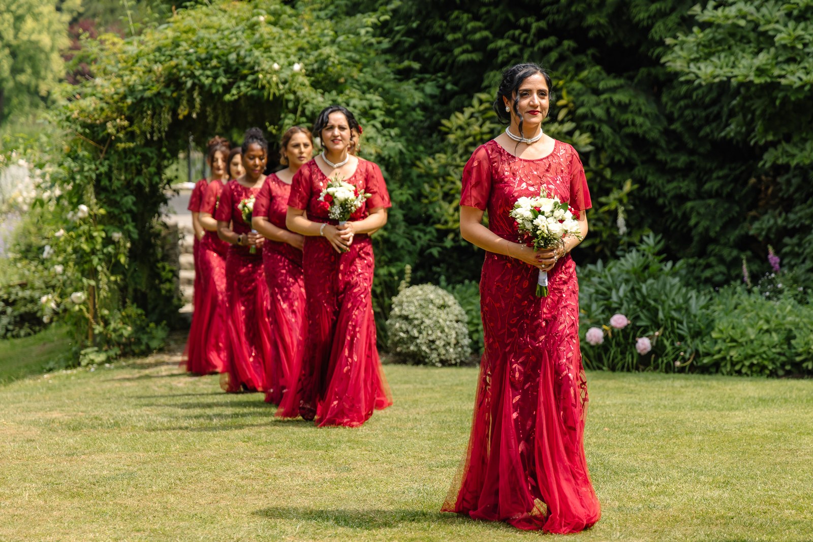 Bridesmaids in red carry bouquets down the garden aisle during the processional