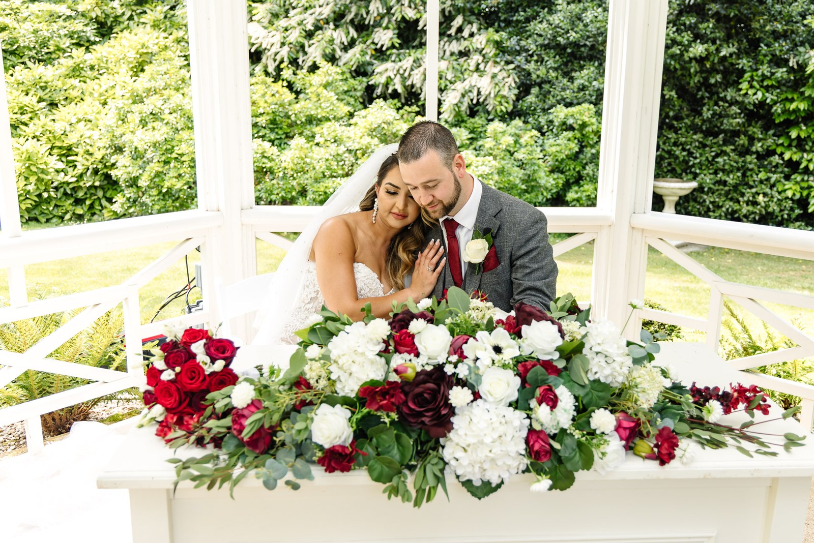 The couple lean close in the white garden pavilion above a red and white floral display