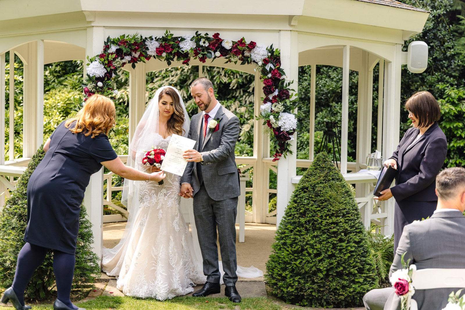 Bride and groom hold their marriage certificate in the pavilion beside a red floral arch