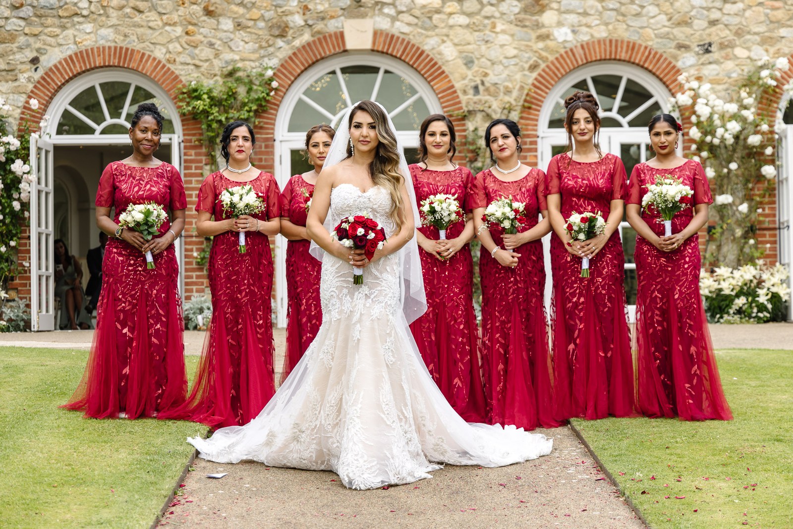 Bride with her red-gowned bridesmaids before the flint-and-brick arches of The Orangery
