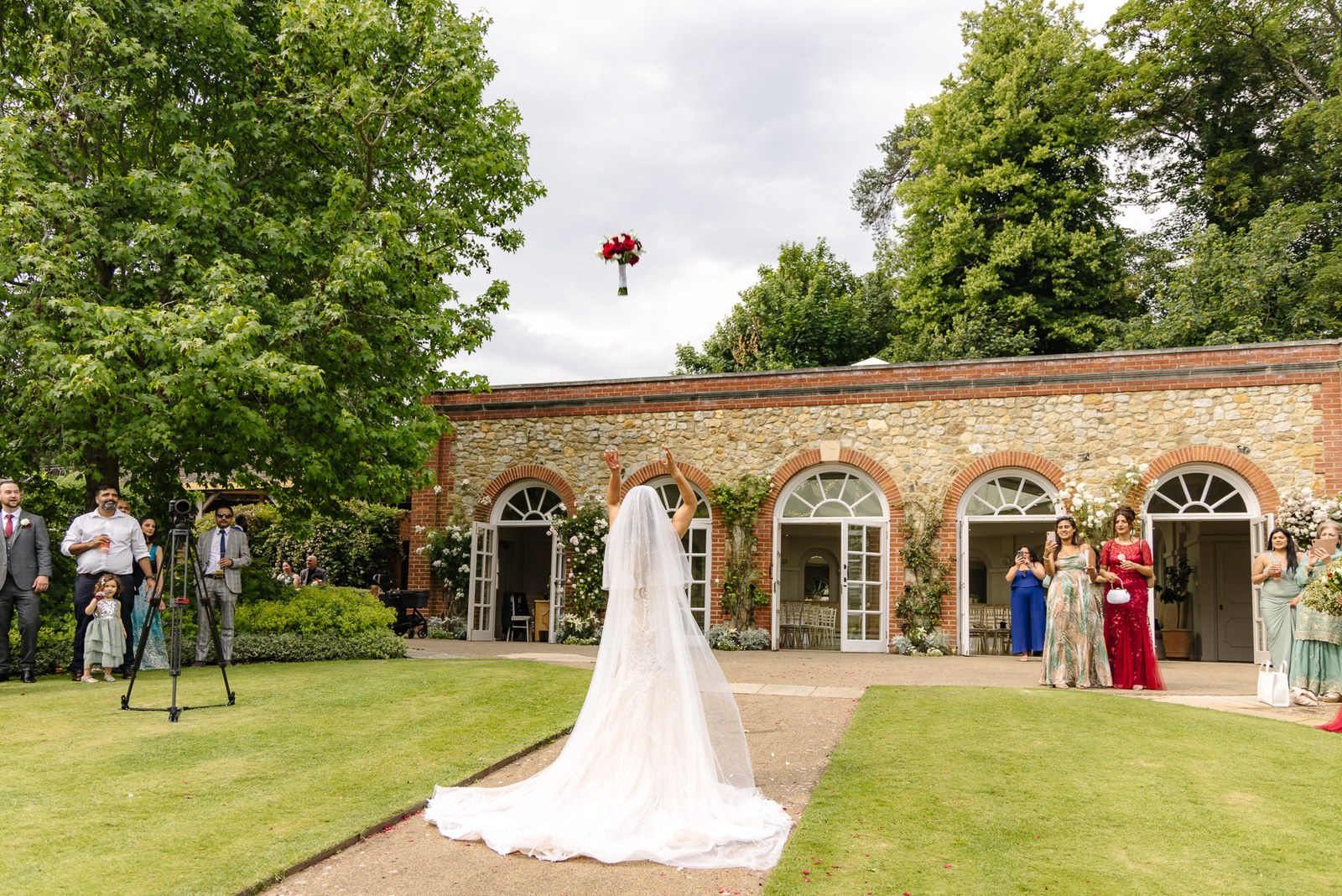 The bride's long train trails on the lawn as she faces The Orangery