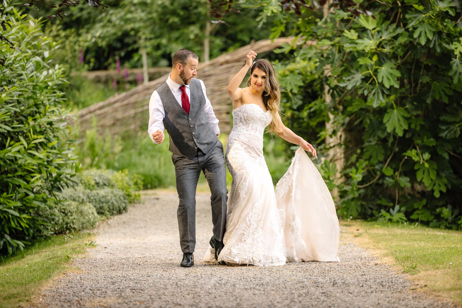 The groom leads the bride along a gravel garden path as she lifts her train