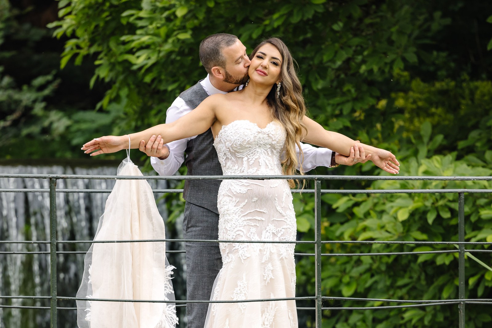 The groom kisses the bride as she leans back on the railing above the water