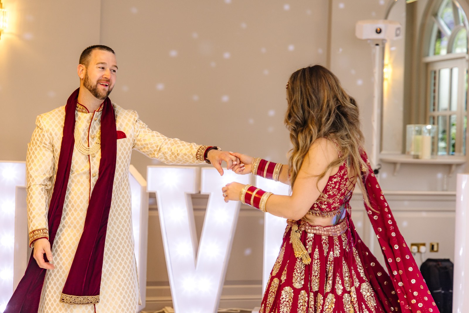 Bride and groom dance before illuminated light-up letters, the groom in a cream sherwani