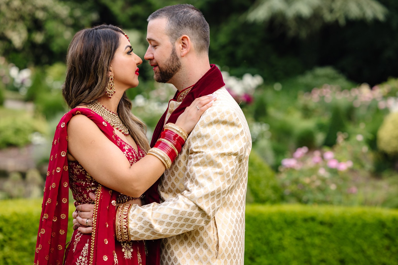 The couple embrace in the gardens in red and gold Indian attire