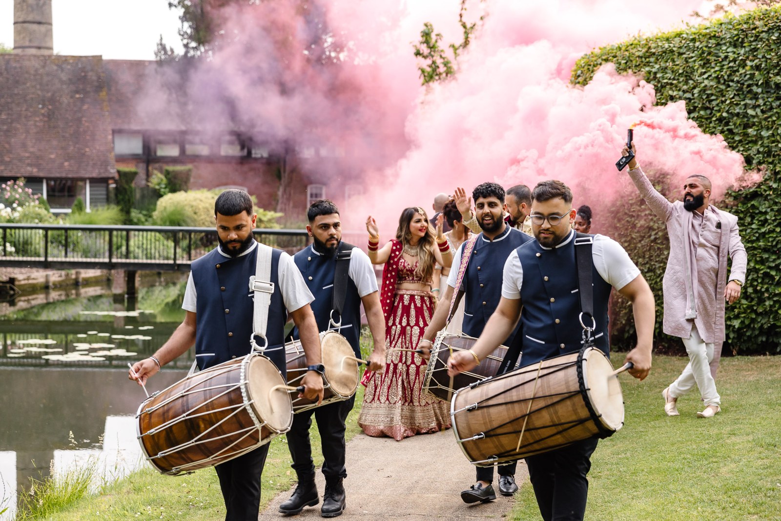 Dhol drummers lead the bride along the path beside the lake through pink smoke