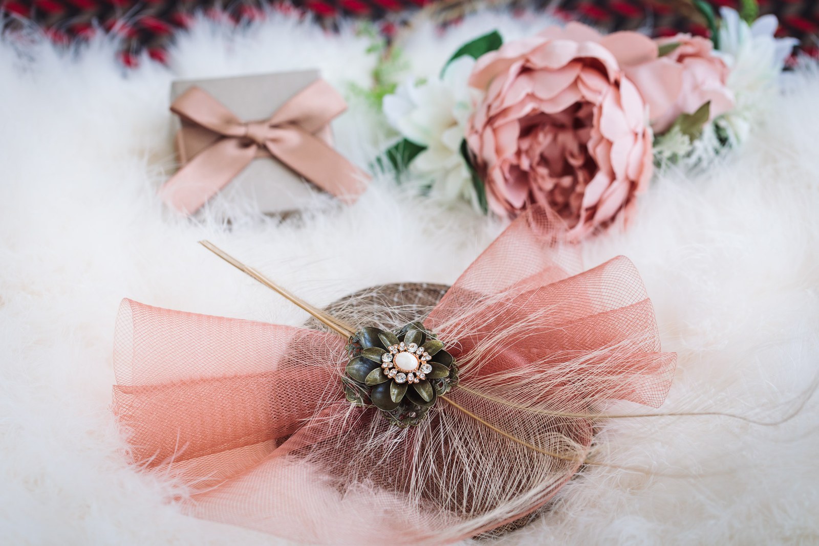Flat lay of a dusky pink fascinator, silk peony and ring box on white fur