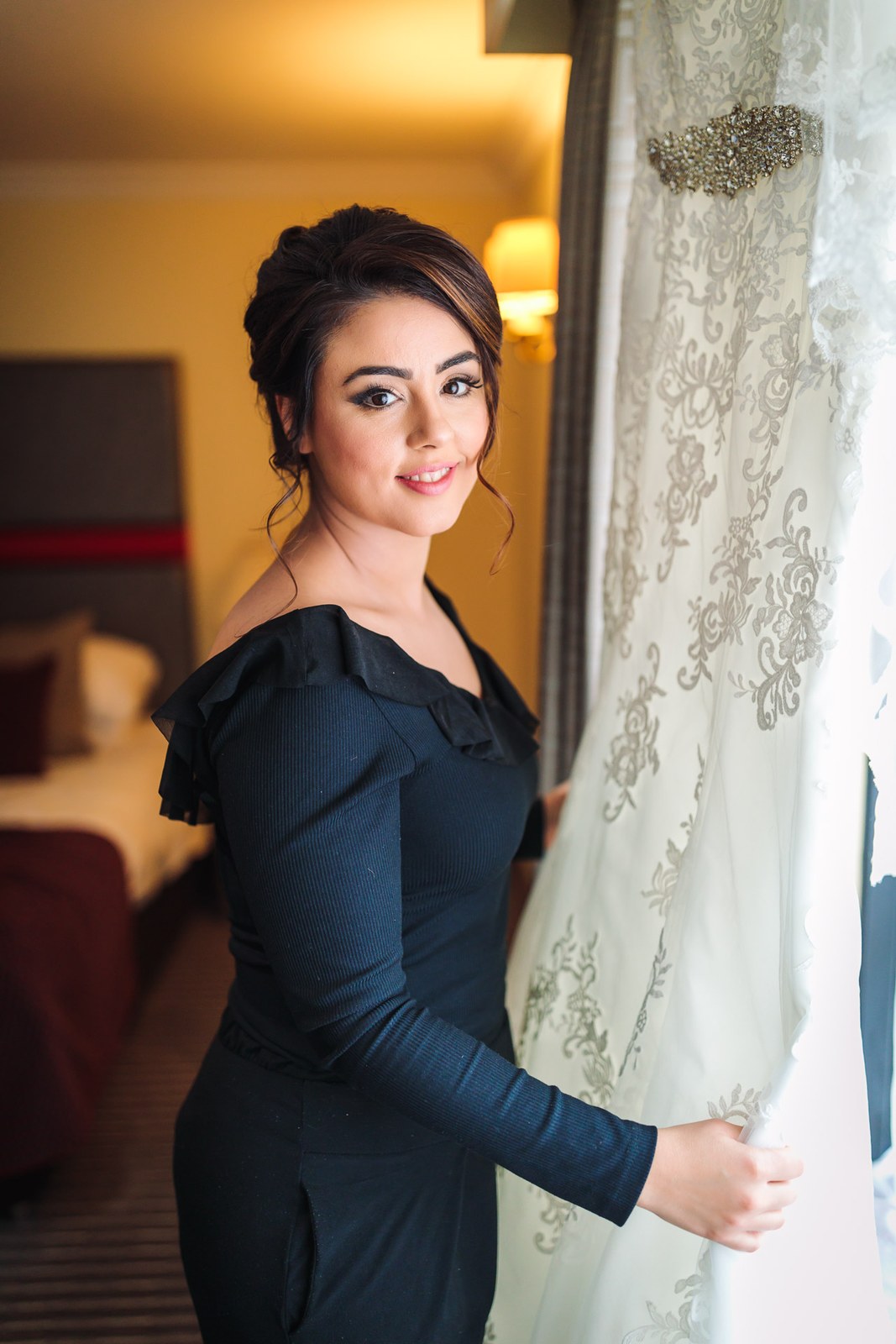 The bride in her black off-shoulder top beside the gown hanging at the window