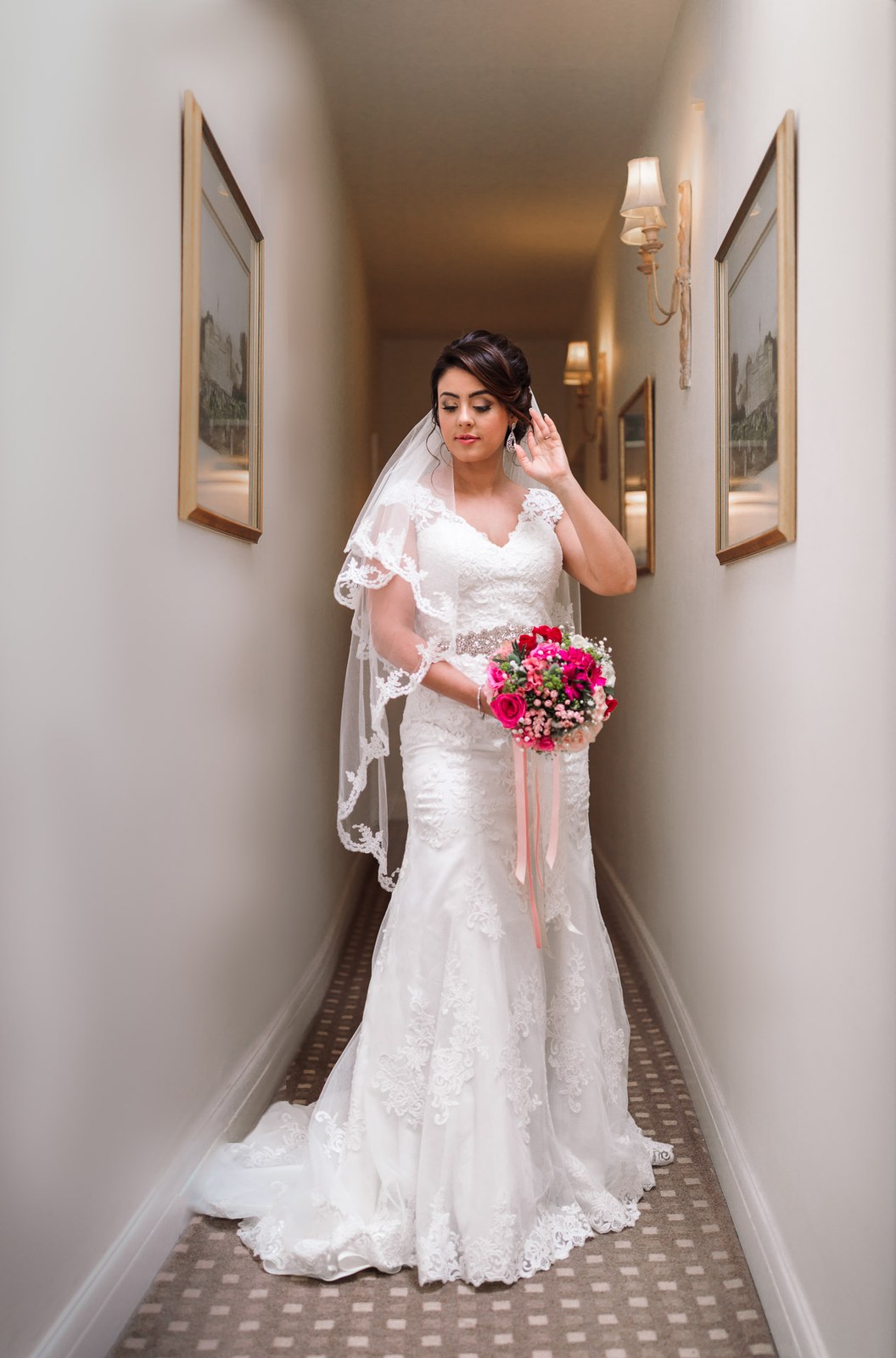 The bride full length in a picture-hung hotel corridor, touching her earring