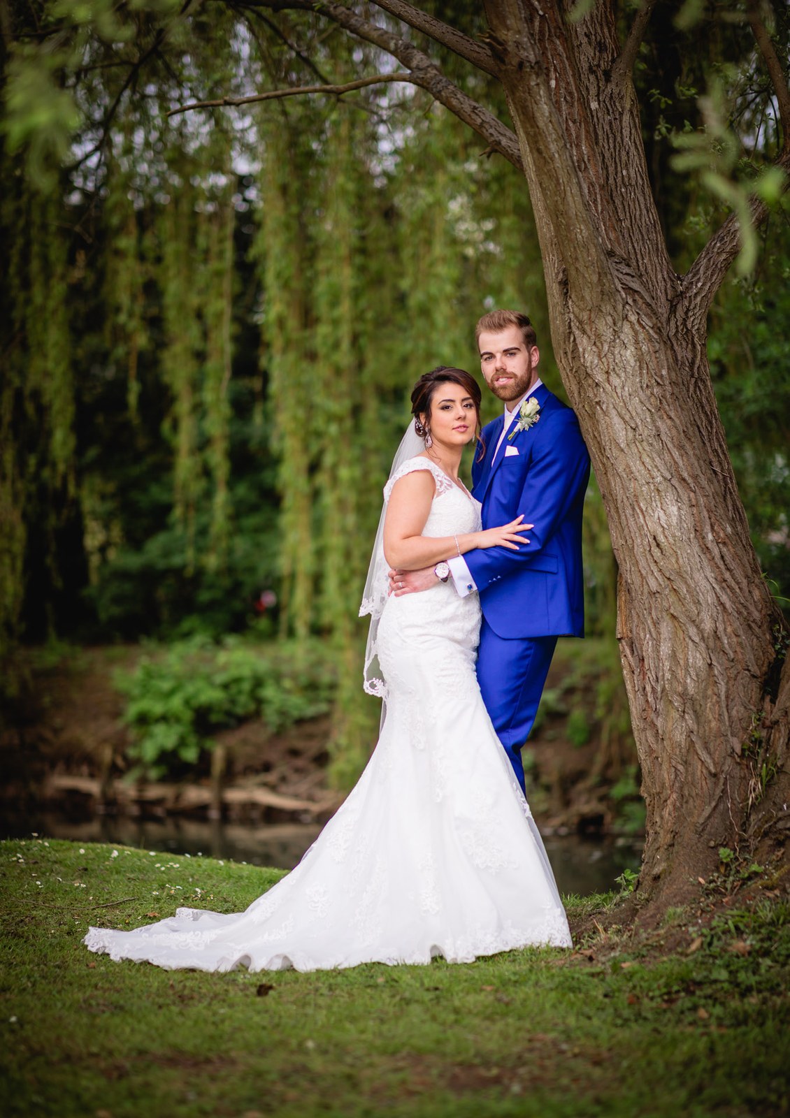Bride and groom embracing under a riverside willow, her train on the grass