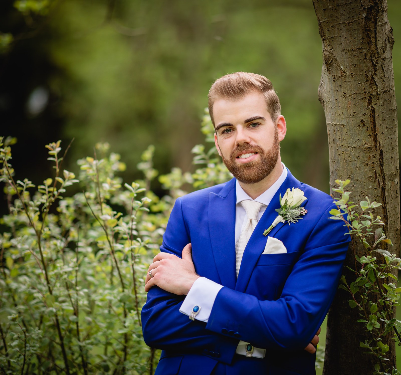 Portrait of the groom in a royal blue suit, arms folded, against a tree