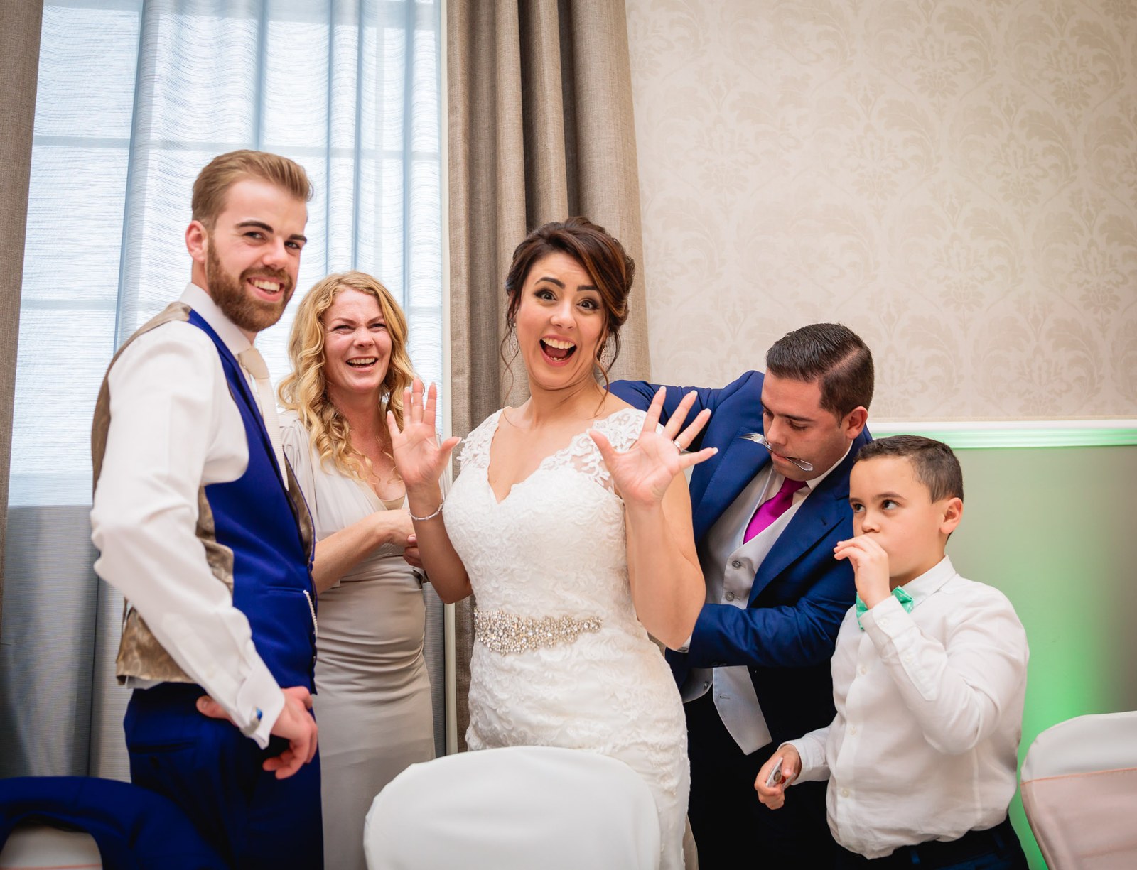 The bride pulling a face with her hands raised as the groom and guests laugh at the reception