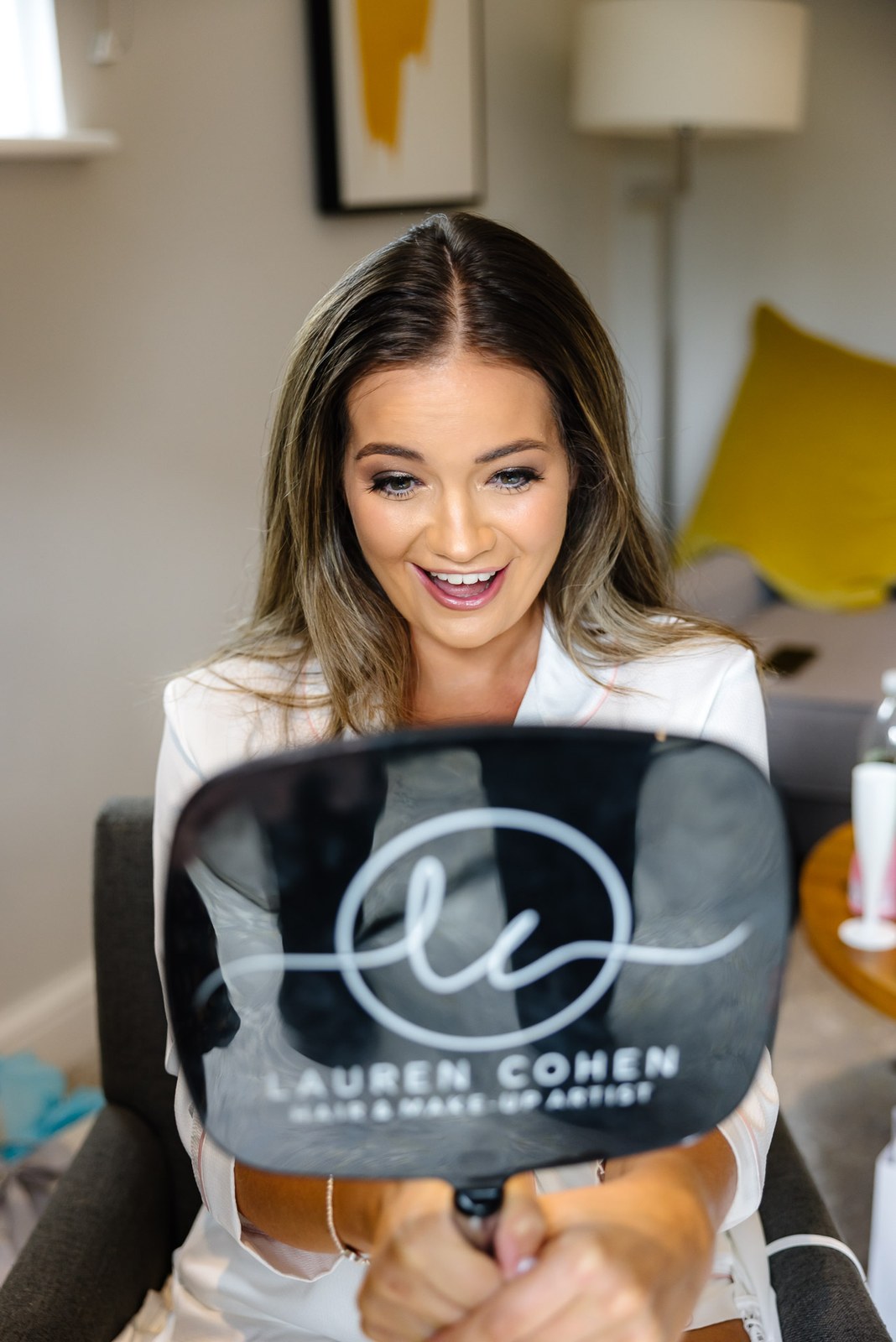 Bride laughing as she checks her make-up in a branded hand mirror in the getting-ready room
