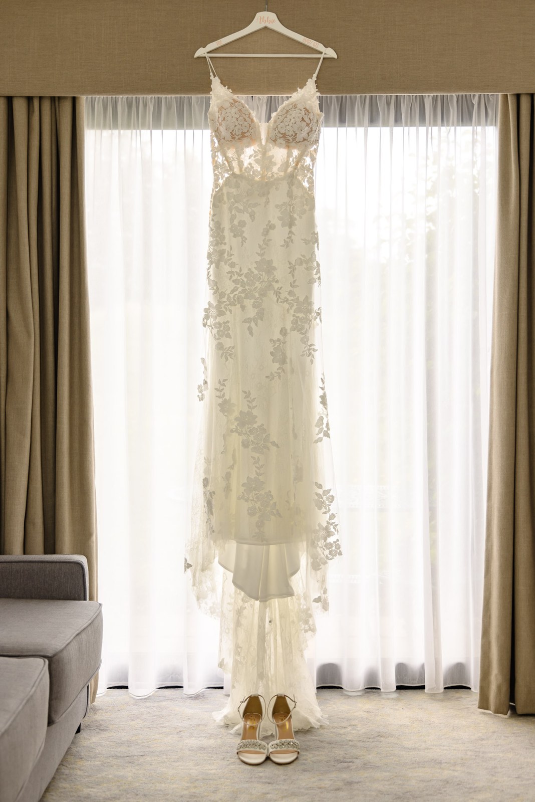 The lace wedding gown hanging by the window with bridal shoes below
