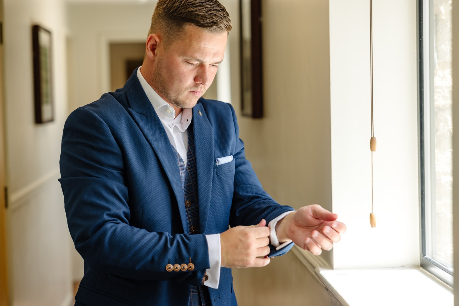 Groom fastening his watch by a window in his navy suit
