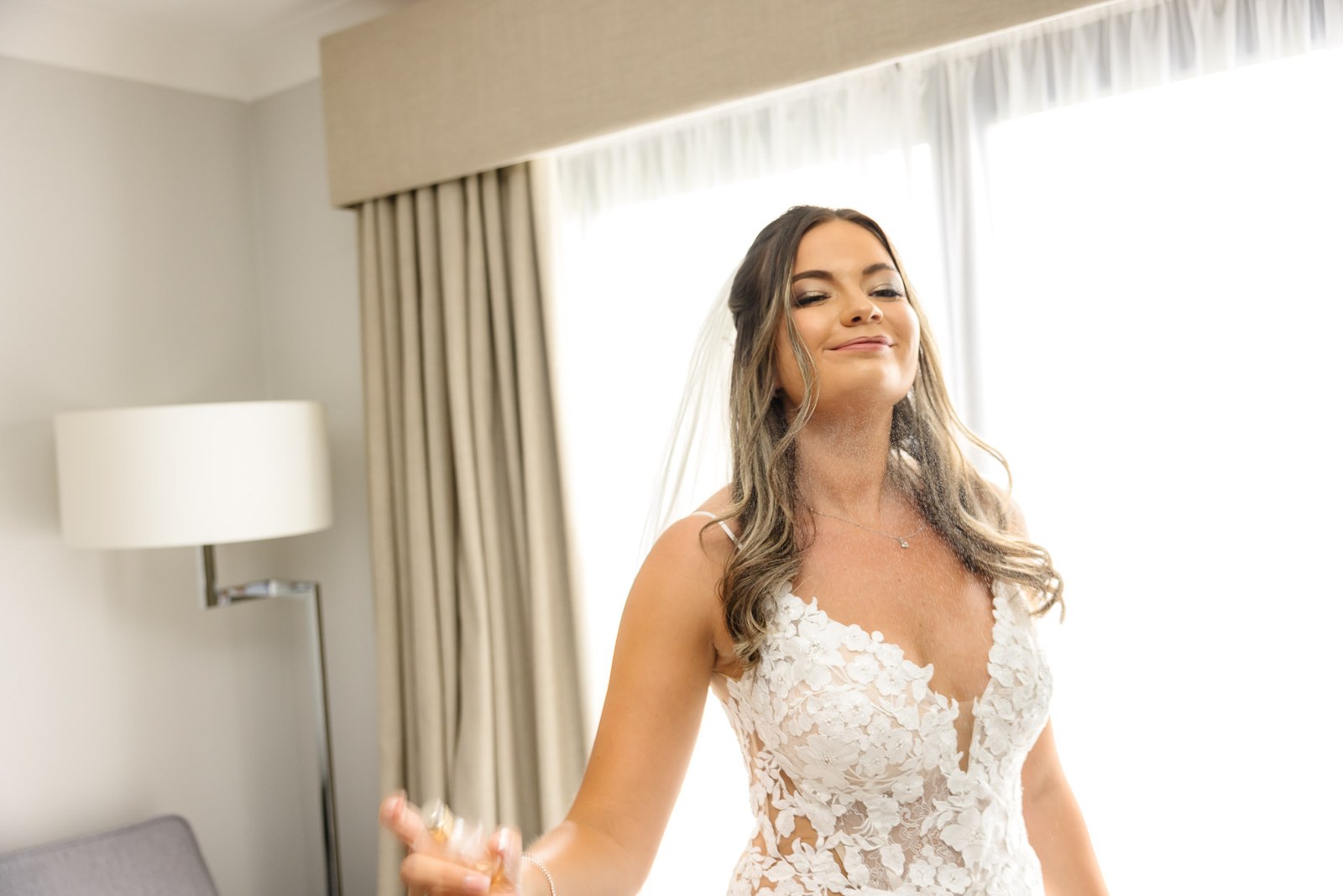 Bride opening her arms in her lace gown and veil beside the window