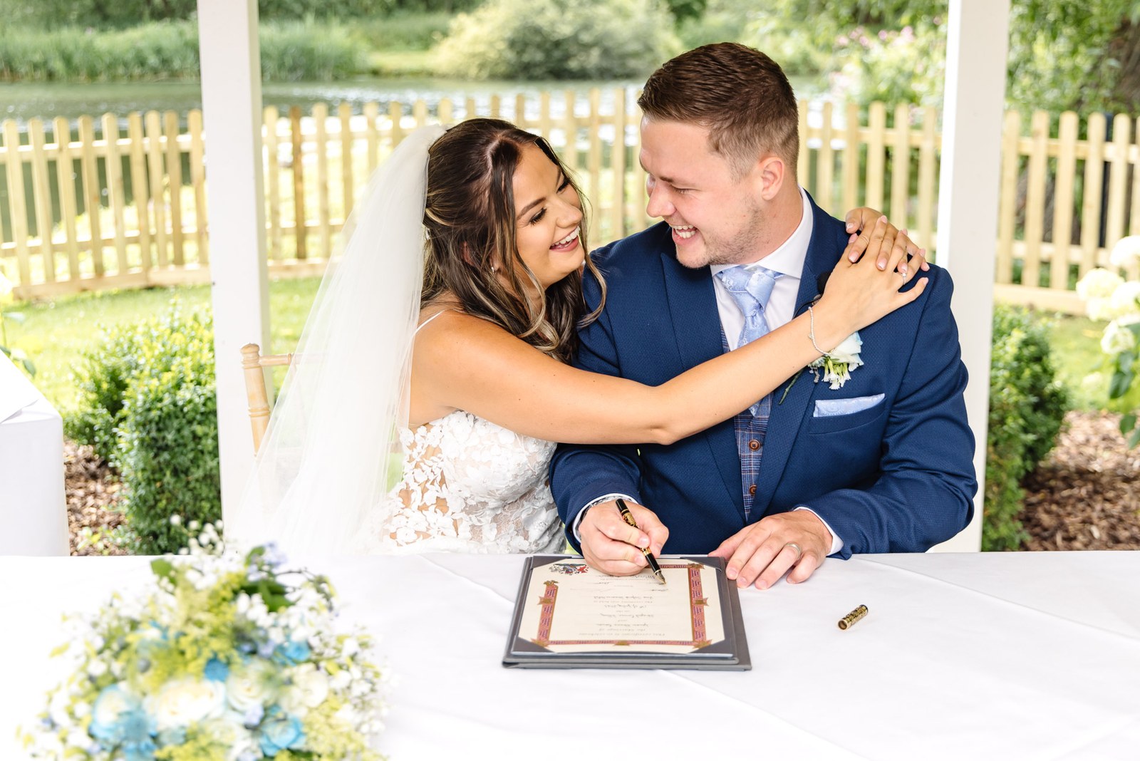 Bride embracing the groom as he signs the register beside the river