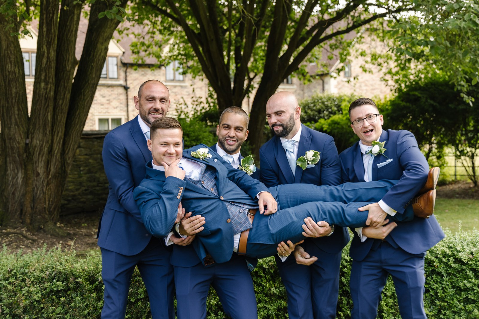 Groomsmen lifting the laughing groom on the lawn at voco Oxford Thames