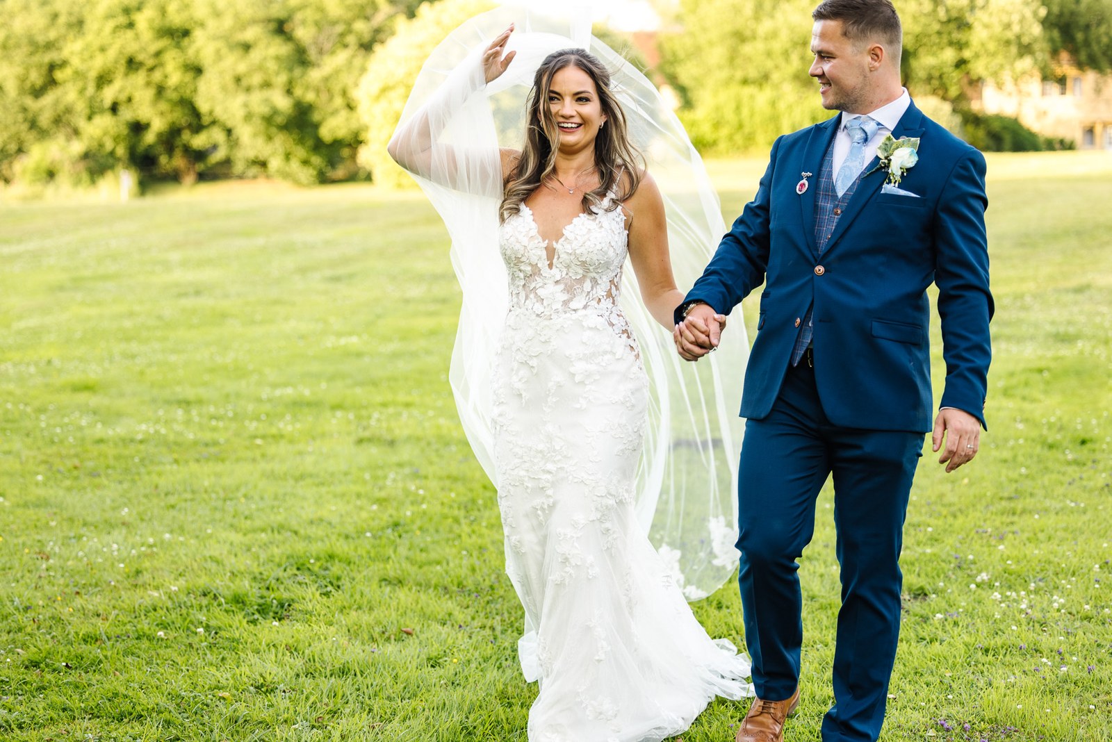 The couple walking hand in hand across the sunlit lawn, veil lifted