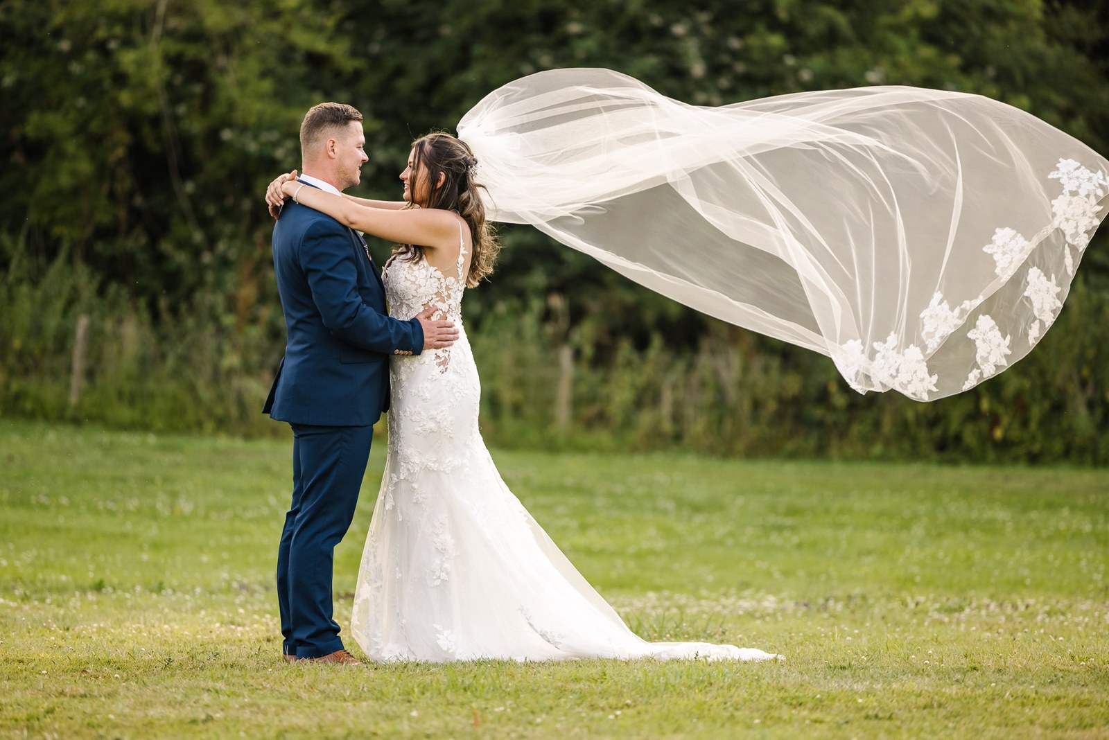 Bride and groom embracing as her veil streams in the summer breeze