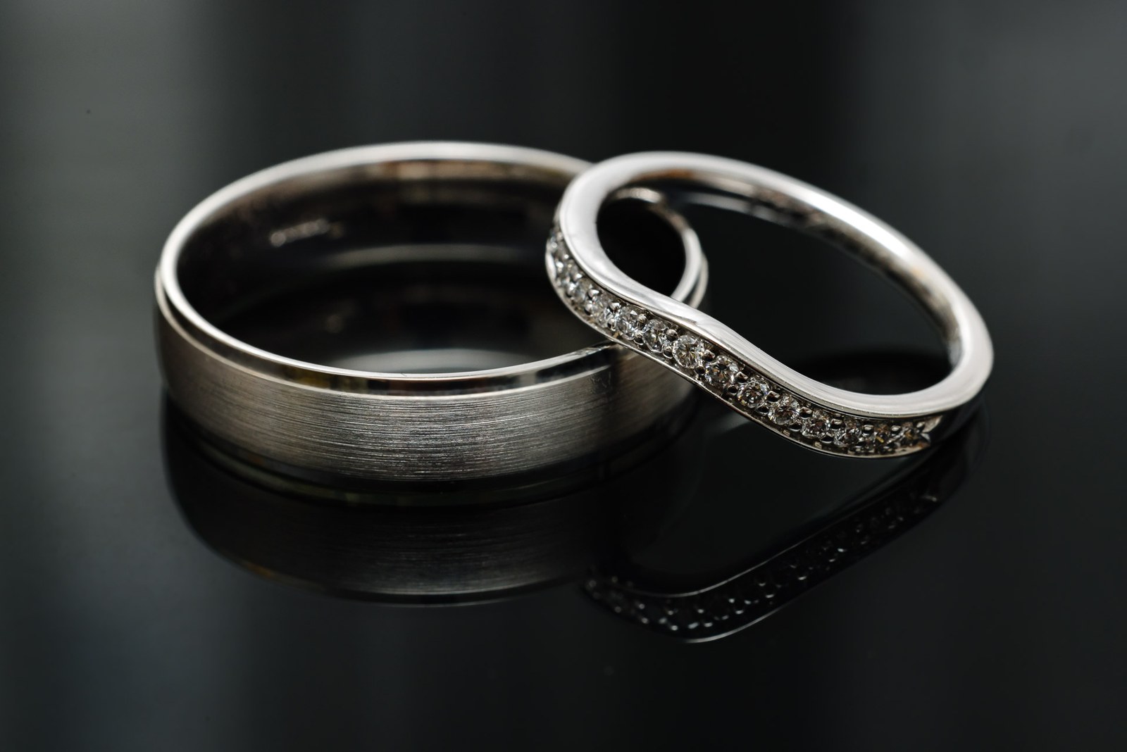 Two wedding rings, one brushed band and one diamante band, on a dark reflective surface