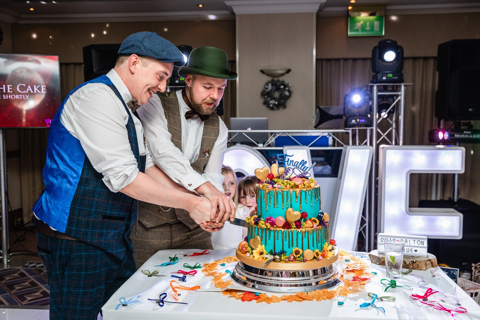 The grooms cutting their teal drip cake beside illuminated love letters