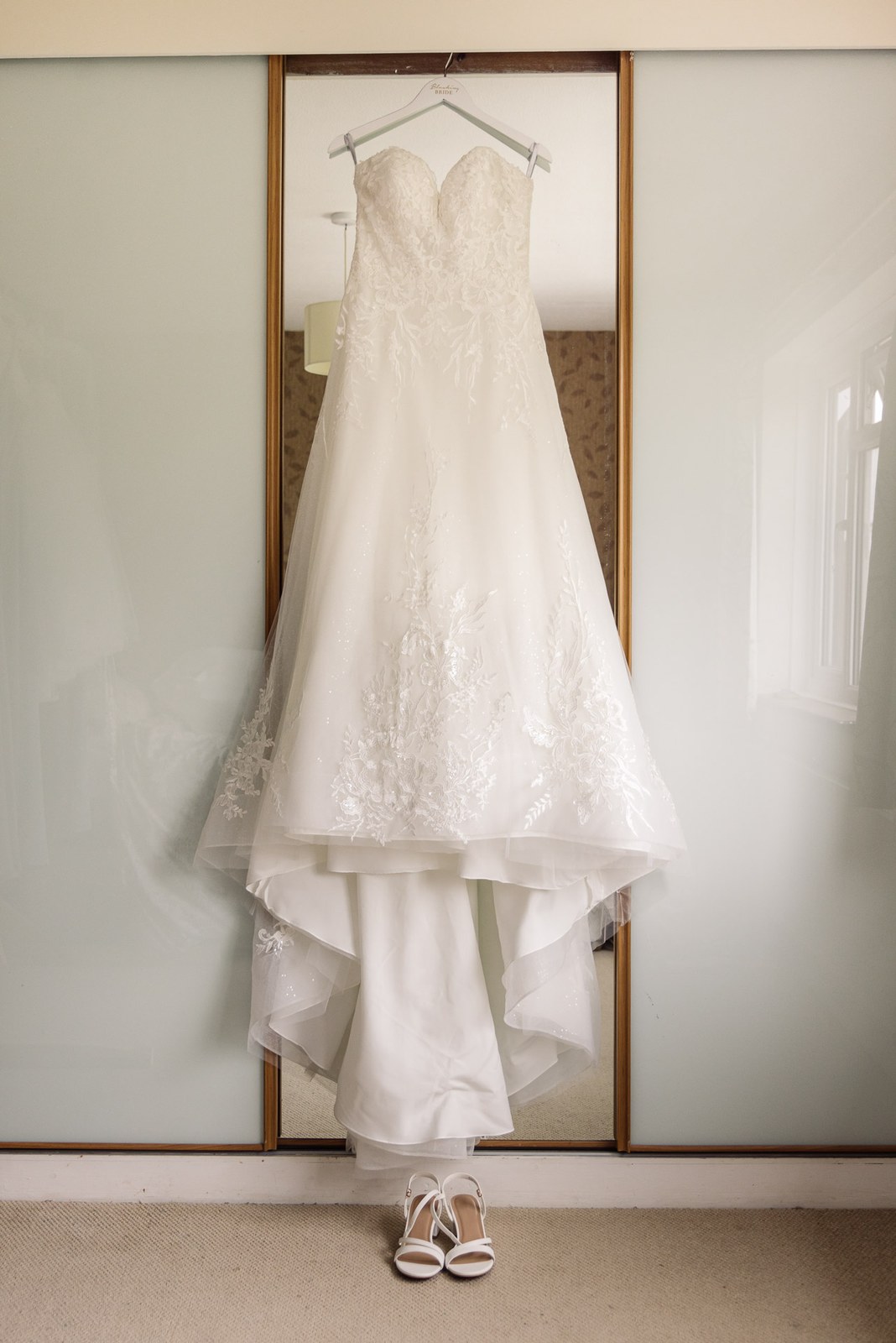 The lace wedding gown hanging against a mirrored wardrobe with shoes below