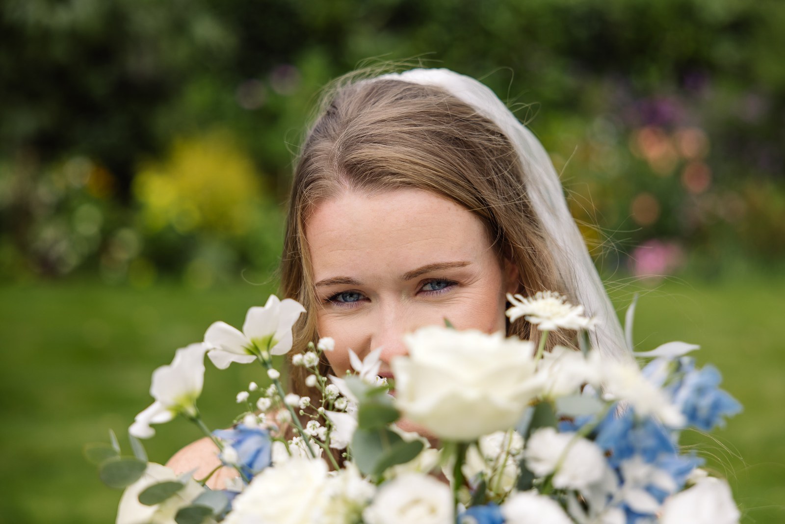 Bride peeking playfully over her white and blue bouquet in the garden
