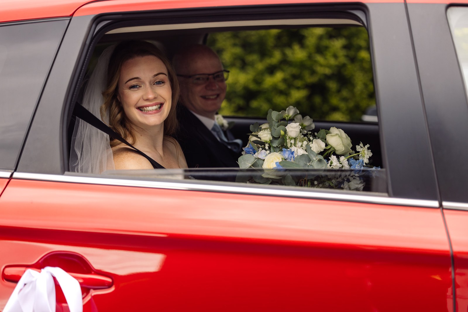 Bride arriving with her father in a red car, her bouquet resting on the window ledge
