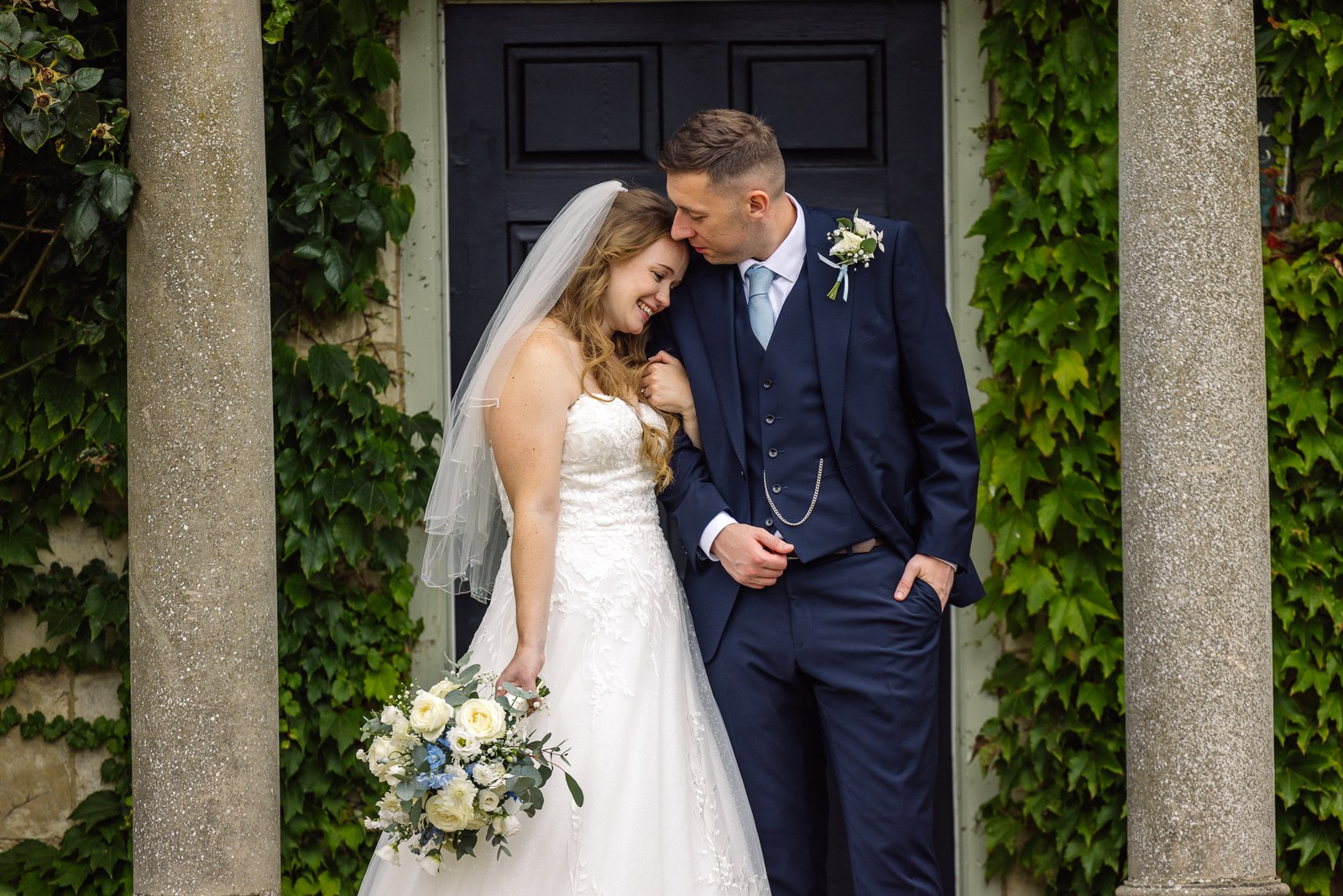 The couple at the ivy-clad doorway as the groom kisses the top of her head