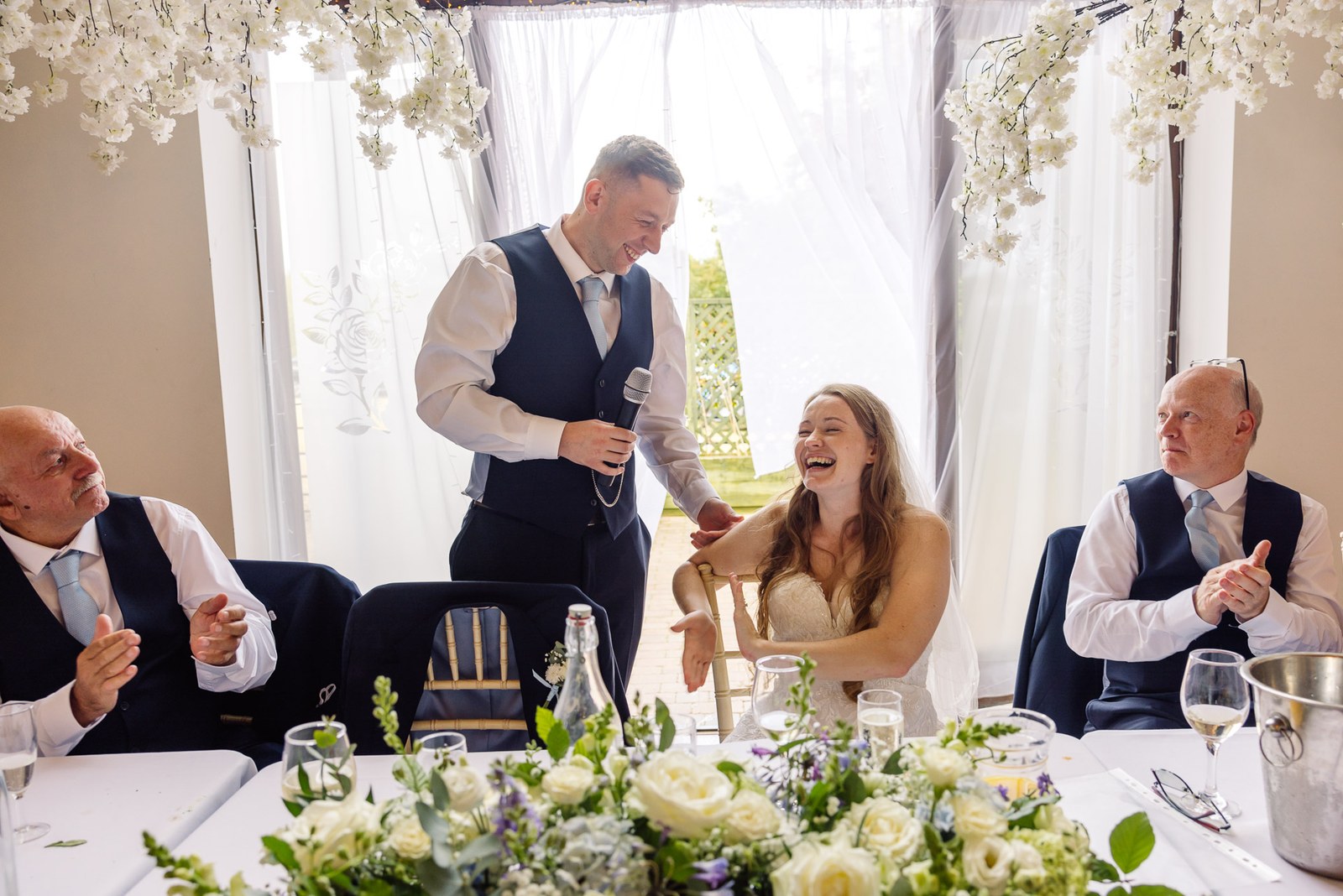 The groom's speech at the top table as the bride laughs beneath a floral backdrop