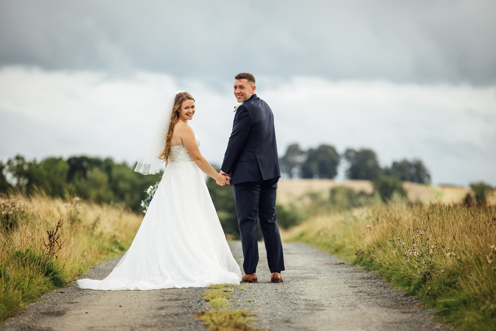 The couple holding hands on a country lane beneath a dramatic sky