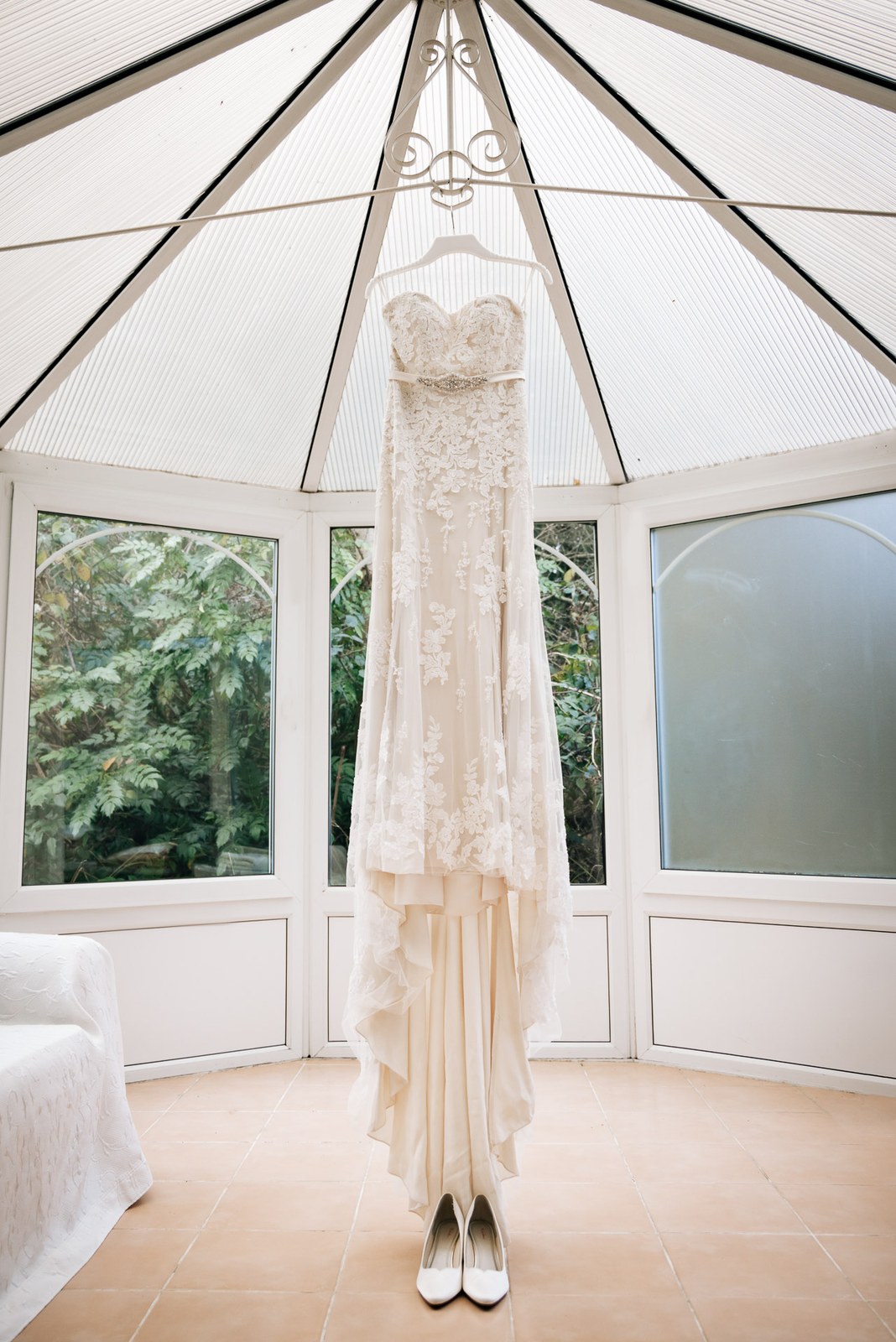 The lace wedding gown hanging in the light-filled conservatory with shoes below
