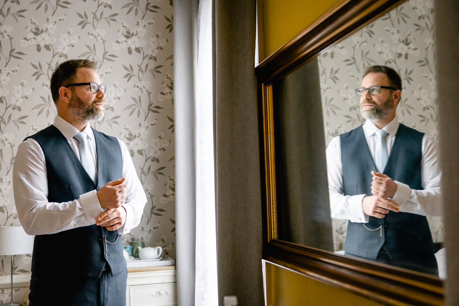 Groom adjusting his suit, reflected in a dark wooden mirror on the floral wallpaper