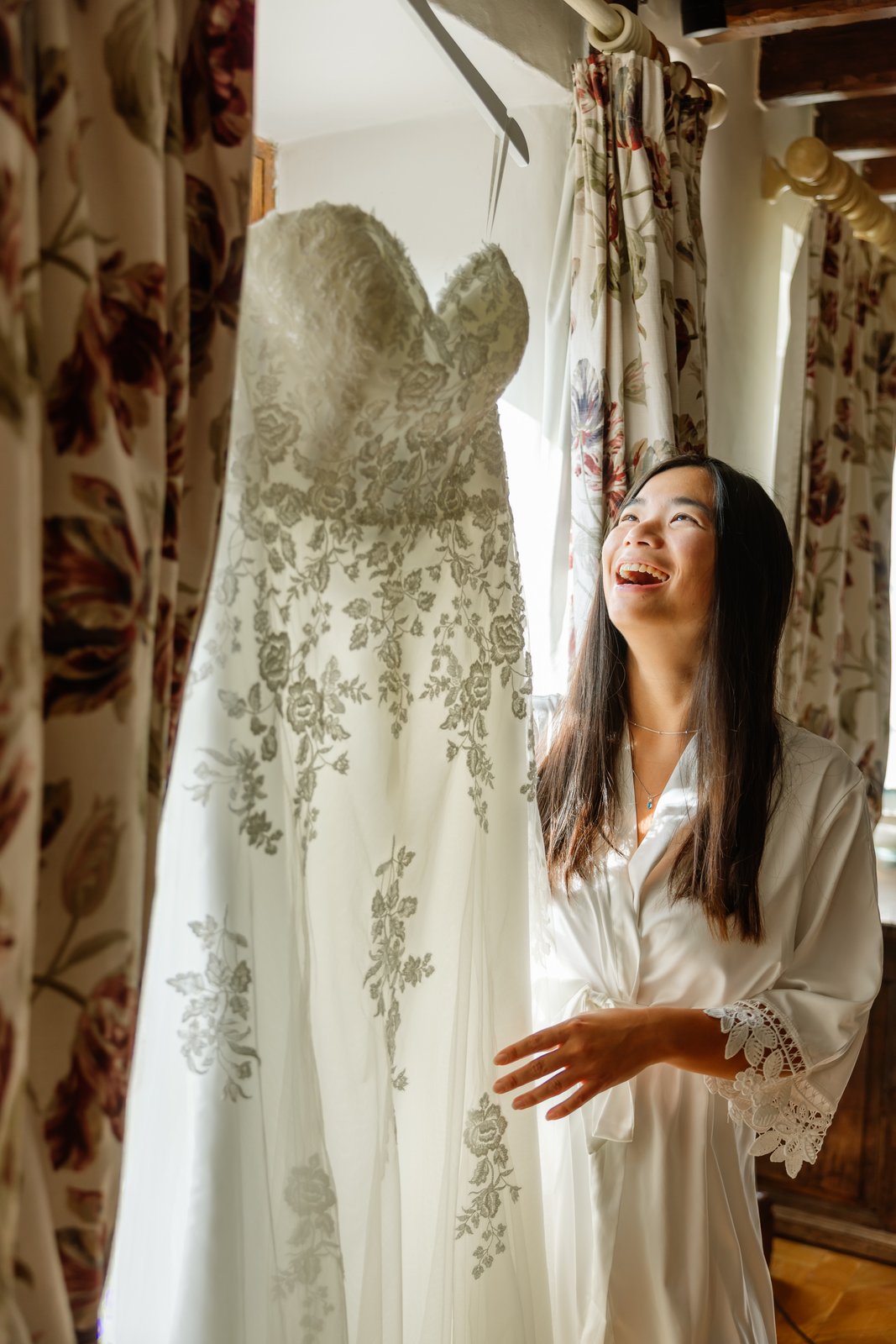 Bride in a silk robe laughing beside her lace wedding dress hanging in a window at Worton Hall.