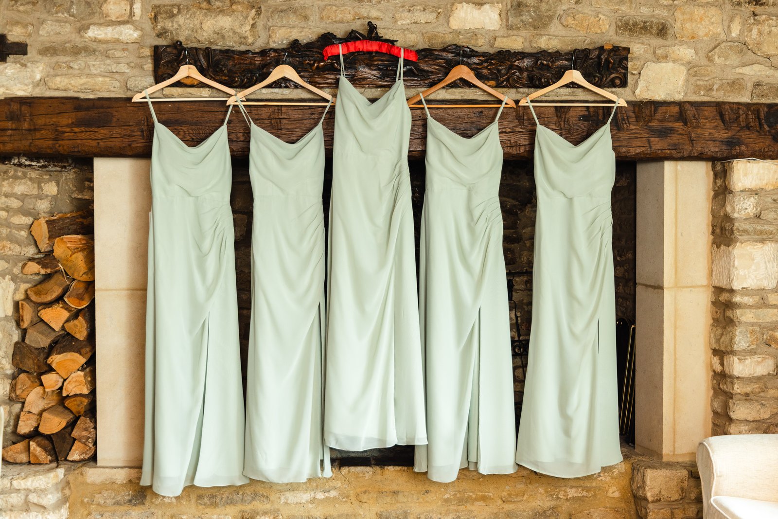 Five sage green bridesmaid dresses on wooden hangers above a Cotswold stone fireplace and log store.
