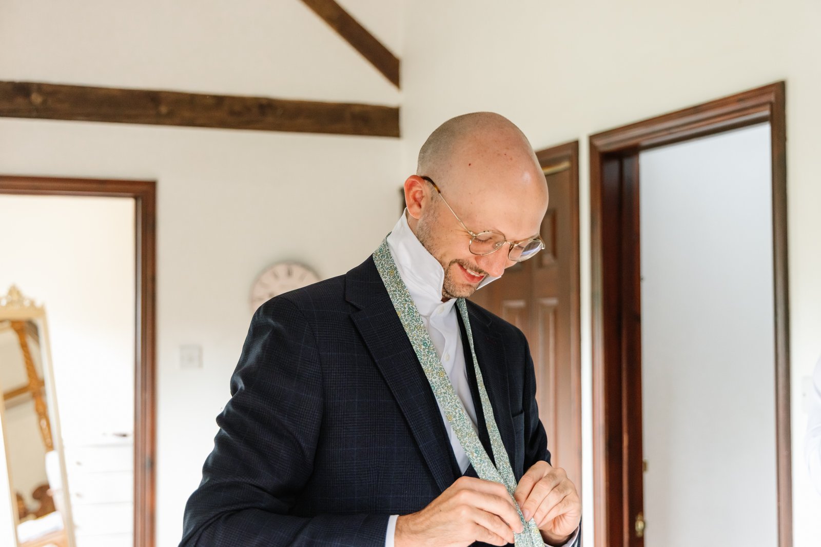 Groom fastening his patterned tie in a beamed dressing room before the wedding.
