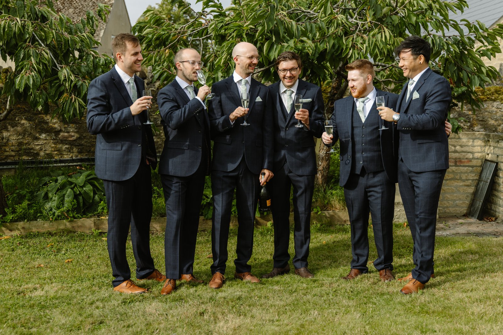 Groom and his groomsmen in navy suits sharing a drink on the lawn at Worton Hall.