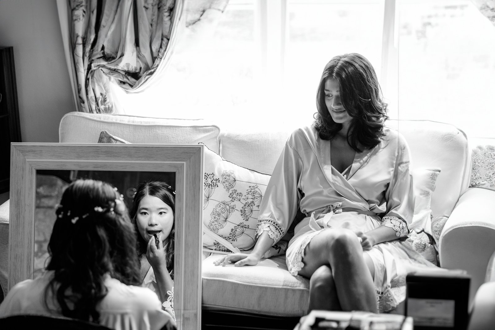 Black and white portrait of the bride putting on lipstick at a mirror, a bridesmaid behind her.