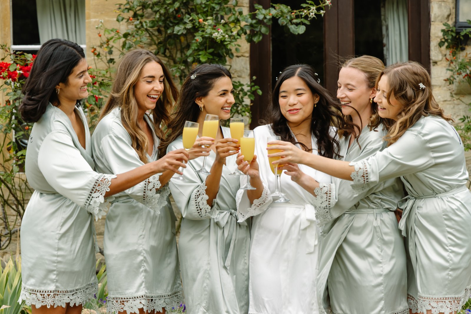 Bride and bridesmaids in matching robes toasting together outside a stone cottage.