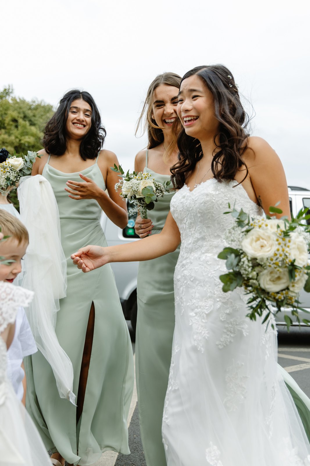 Bride walking with her bridesmaids, one carrying her veil, another a white and green bouquet.