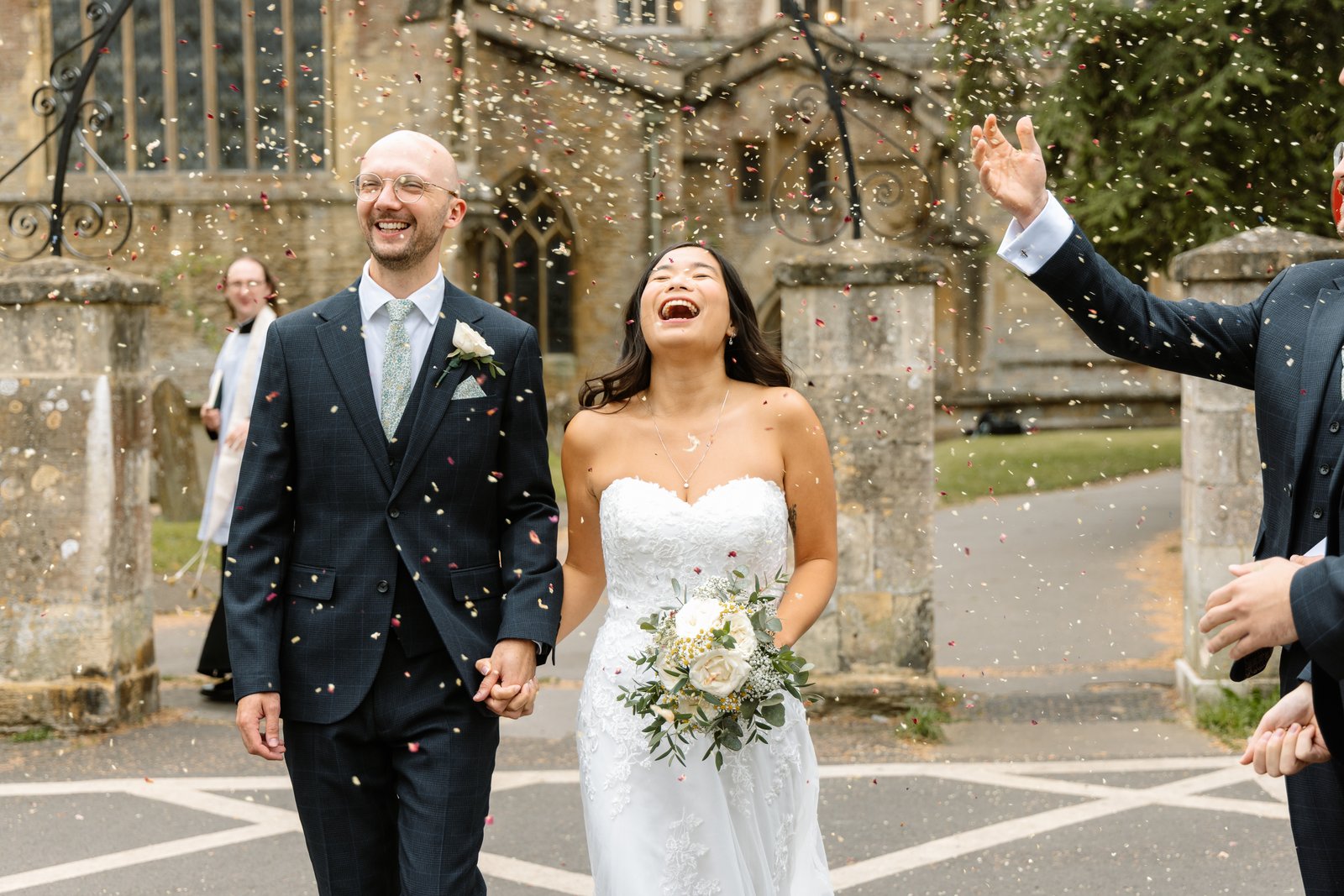Bride and groom laughing as confetti falls over them outside the church gateway.