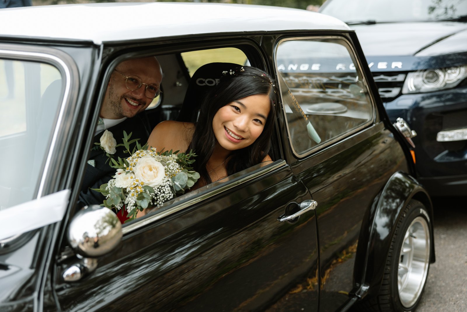 Bride leaning out of the window of a black classic Mini decorated with ribbon.