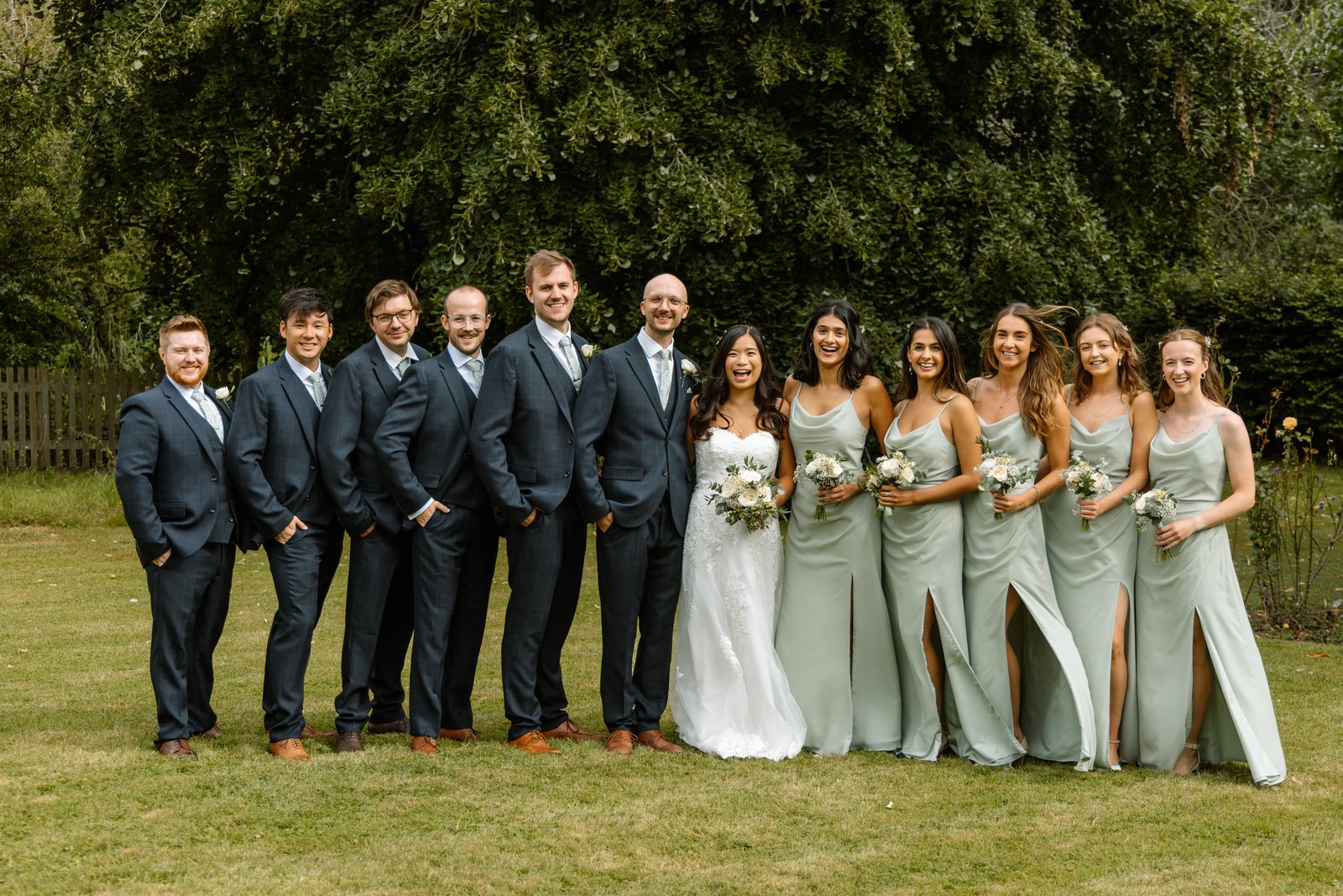 The full wedding party lined up on the lawn at Worton Hall in front of a tall hedge.