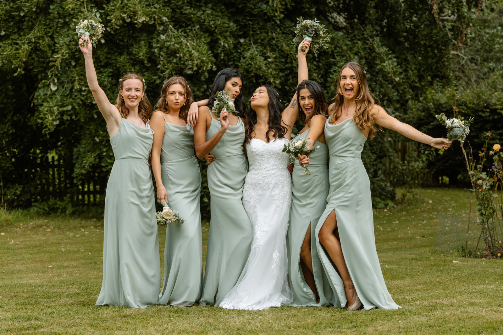 Bridesmaids cheering with raised bouquets around the bride on the lawn.