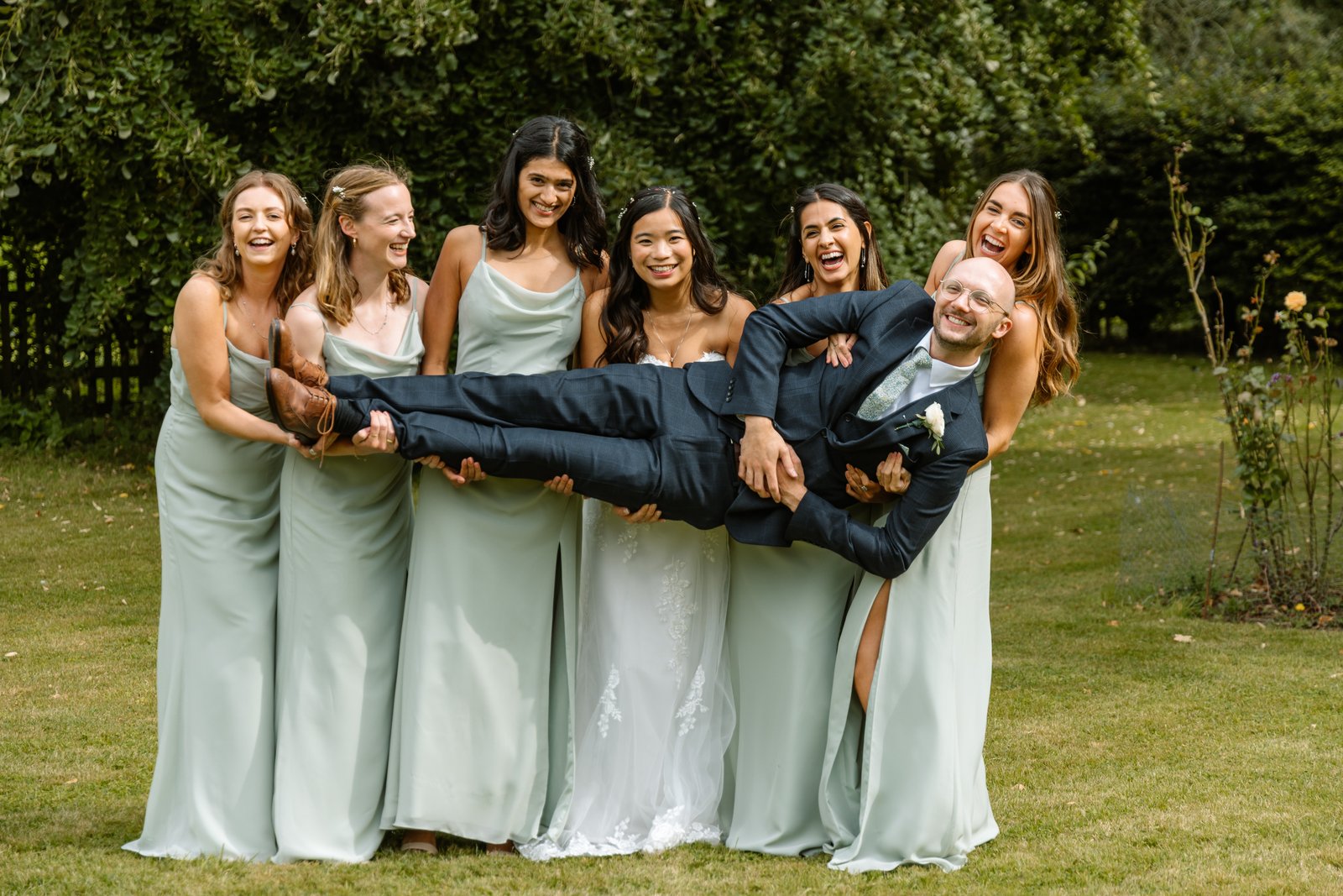 Bridesmaids carrying the groom horizontally while the bride laughs beside them.