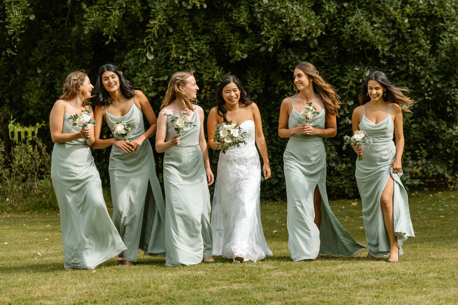 Bride walking across the grass with her bridesmaids in sage green dresses.