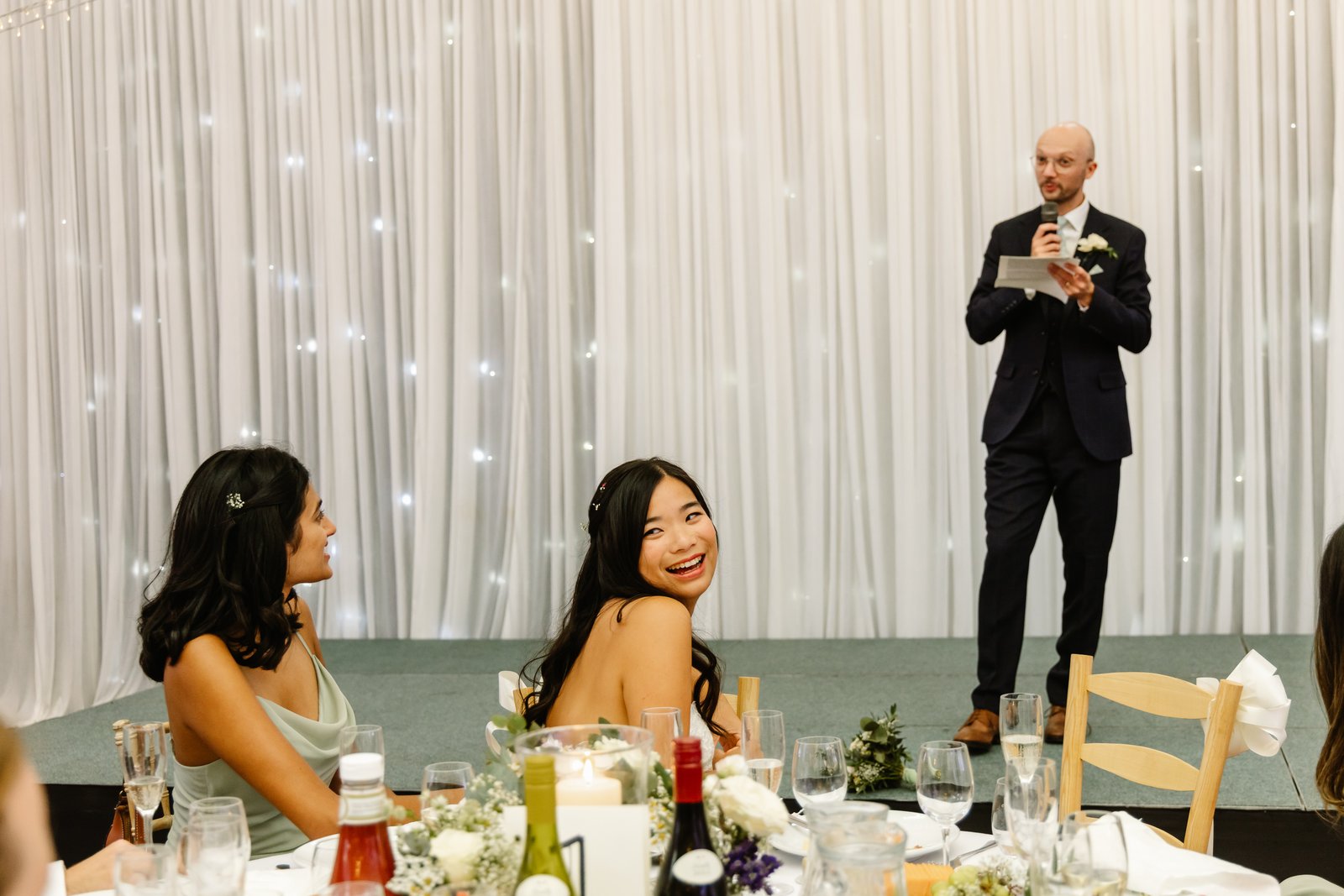 Groom giving his speech in front of a draped fairy-lit backdrop as guests listen.