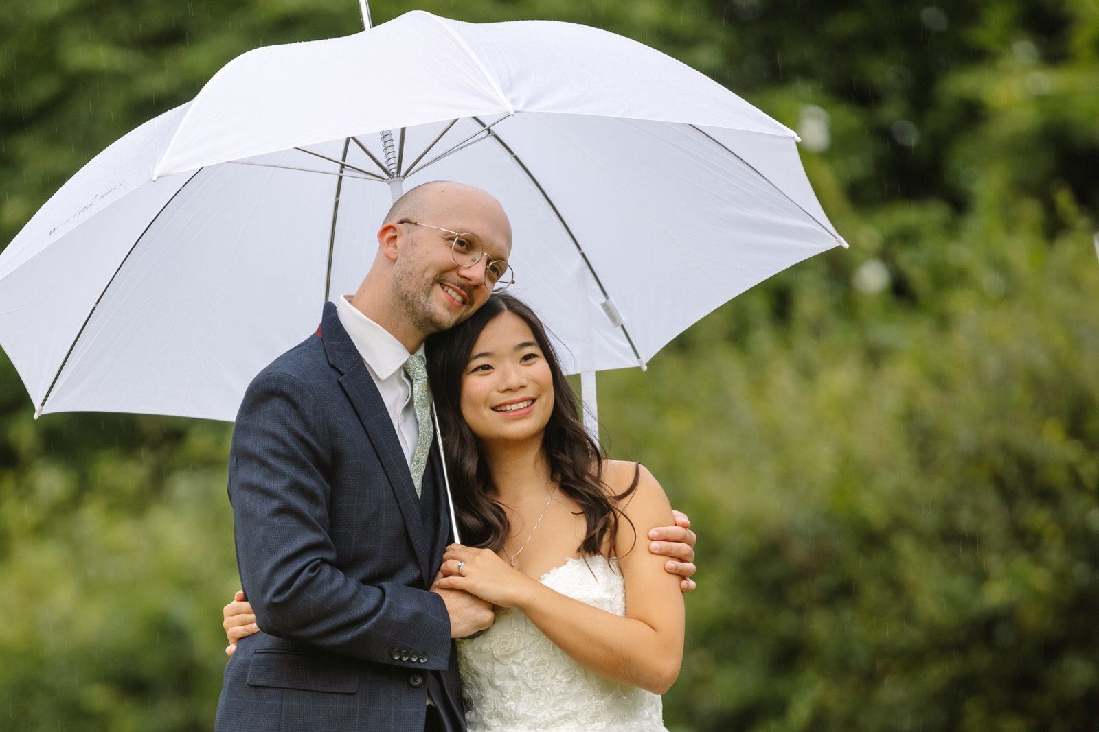Bride and groom sheltering close together under a white umbrella in the gardens.