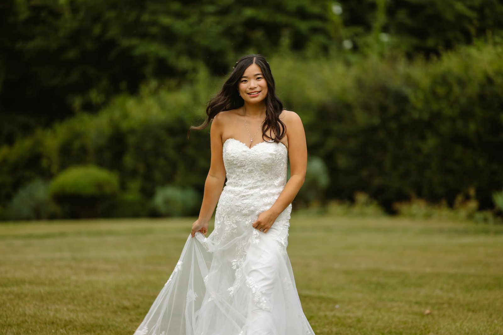 Bride standing on the lawn holding her dress, with hedges and trees behind her.